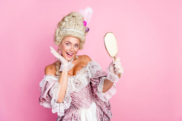 Elegant woman in baroque costume admiring herself in handheld mirror with a pink background