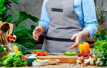Chef holds hands over cutting board with chopped paprika. Cozy kitchen with wooden table, kitchenware, vegetables, herbs and ingredients for cooking. Healthy vegan eating, culinary, recipes, food 