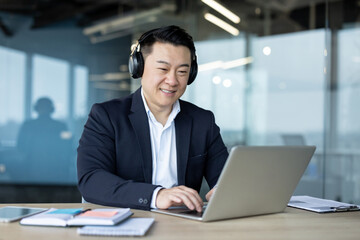 Fototapeta premium Asian young male businessman sitting in the office wearing headphones and smiling while working on a laptop