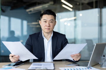 Serious young Asian male businessman working in the office at his desk, holding documents and a contract in his hands, spreading his arms in frustration and looking at the camera