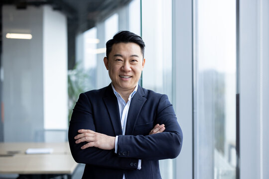 Portrait of a serious young Asian man in a business suit confidently posing for the camera in a corporate office with his arms crossed on his chest