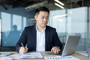 Serious and focused young Asian man sitting at a desk in the office and working on a laptop, making...