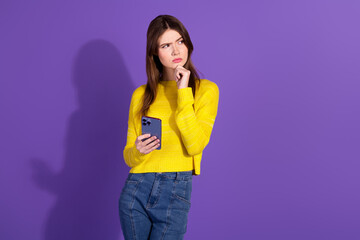 Stylish Young Woman Holding Smartphone Posing Against Purple Background in a Casual Yellow Sweater and Trendy Jeans