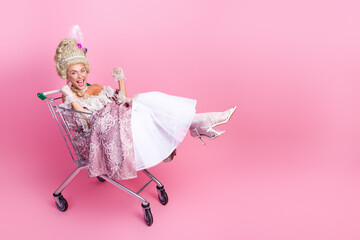 Playful woman dressed as a royal figure with an elegant costume riding in a shopping cart against a pink background