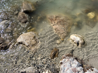 riverbank with assorted rocks and sand. intricate natural patterns. soft water movement. water surface. water reflection. sunlight pattern. nature abstract