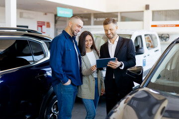 Professional car salesman in black suit showing details on tablet to smiling young couple in modern...