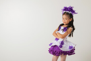 Adorable girl dressed as a cheerleader in a white and purple layered outfit, accessorized with a purple feather headpiece. Arms crossed, studio portrait on an isolated white background