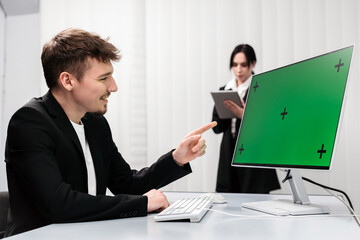 An IT specialist works at a desktop computer with a Chroma Key green screen while a colleague brainstorms in the office. E-commerce concept development, marketing and data analysis in the office.