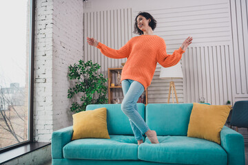Obraz premium Young woman in an orange sweater enjoying her time at home on a teal sofa in a cozy and modern apartment with natural daylight