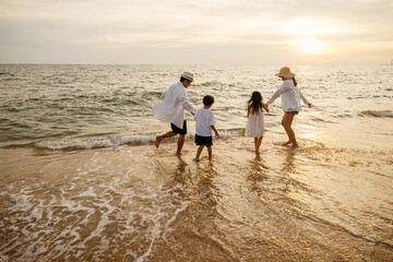 Back view of happy Asian family on beach. Parents and daughters share playful moments running...