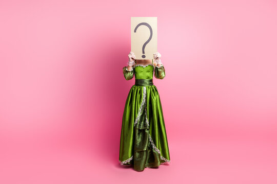 Elegant lady in a green dress hiding face with a question mark sign, against a pink background