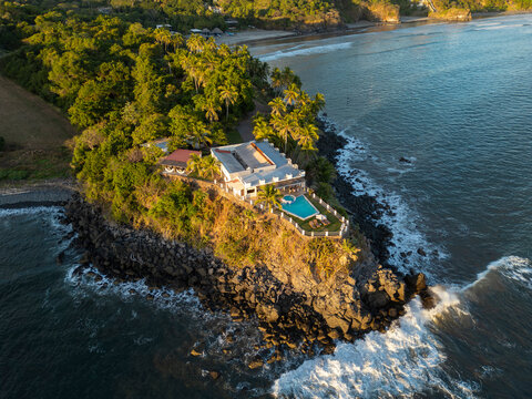 Aerial drone photo of vacation house with pool at Pacific Ocean coast, cliffs and lush green mountains, high waves. Sunny day, blue sky. El Cuco beach, El Salvador. South destinations travel concept.