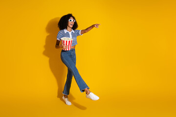 Young trendy girl with curly hair enjoying popcorn and pointing excitedly against a vibrant yellow backdrop