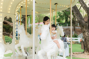Fototapeta premium Mother and child are having fun playing on the carousel,Asian mother and daughter having fun in an amusement park and riding the carousel happily.