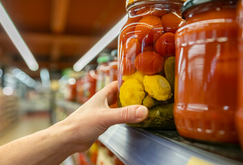 Consumer hand taking assorted vegetables in glass jar from shelf with pickles in supermarket close-up.