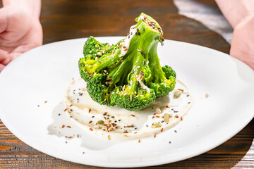 Serving vegetarian dish of steamed green broccoli garnished with sauce and sesame seeds at table.