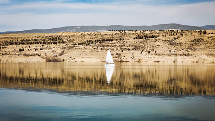 A white sailboat glides across the calm waters of Tbilisi Sea, reflecting the golden hills and blue sky. A peaceful and scenic moment capturing the beauty of nature and relaxation