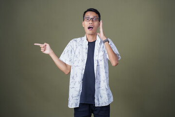 Excited, shocked or excited pose. Shouting with hand pointing to the side of empty space. Asian man with glasses wearing casual white shirt isolated on army green background.