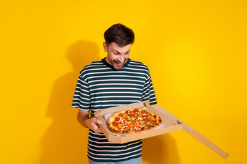 Joyful Young Man in a Striped T-shirt Holding a Pizza Box Against a Yellow Background, Exhibiting...