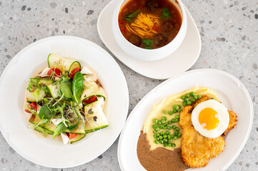 Three delicious dishes on a countertop: soup with noodles and meatballs, zucchini rolls with greens and tomatoes, breaded chicken, mashed potatoes, peas, and sunny-side-up egg.