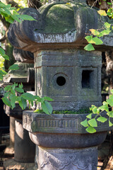 Traditional Japanese stone lantern with detailed engravings shows signs of aging with moss and lichen growth. Sunlight filters through surrounding leaves, casting soft shadows on its textured surface