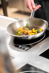 A chef is sautéing fresh vegetables, including Brussels sprouts, yellow bell peppers, and small potatoes, in a stainless steel pan on an induction cooktop in a professional kitchen.