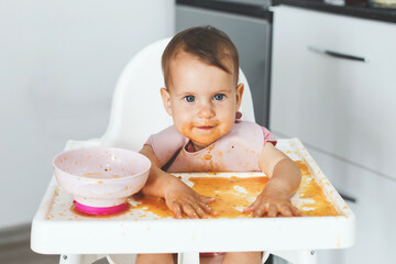 A smiling baby sits in a high chair covered in food, joyfully playing with puree while learning to eat independently in a modern kitchen