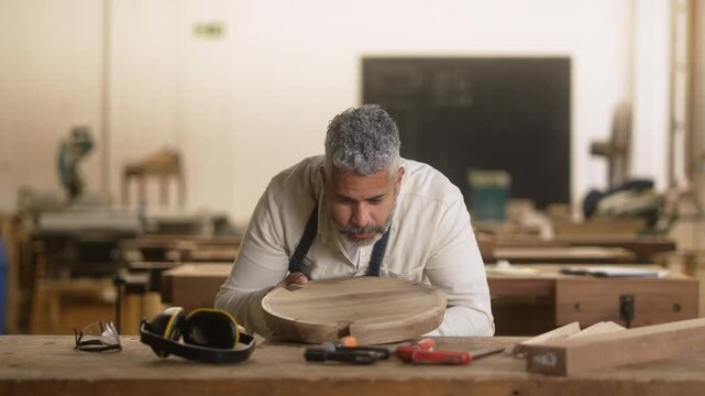 Skilled carpenter blowing dust and admiring his finished woodwork in a workshop. African American man in casual wear carefully examining handcrafted wooden bowl, tools on table
