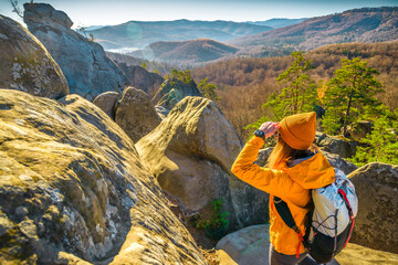 Female hiker wearing an orange jacket, beanie, and backpack stands on a rocky mountain peak,...