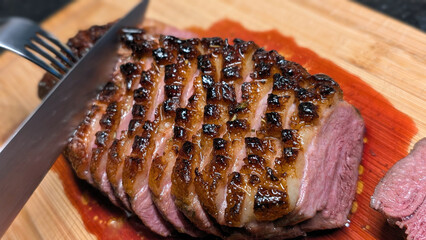 Very close up of slicing medium-rare duck breast with golden crust on a wooden cutting board with a knife and fork in a home kitchen setting. Home cooking concept