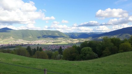 Fototapeta premium Malerische Landschaft mit grünen Hügeln, Dorf im Tal und blauem Himmel mit Wolken, ideal für Natur- und Reisedesigns.