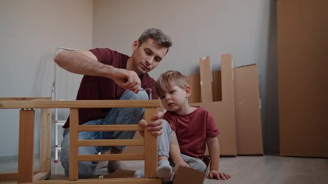 Smiling dad teaches his cheerful little son how to use tools while assembling furniture together at home, creating a happy memory of collaboration and skill-building. Family connection concept.