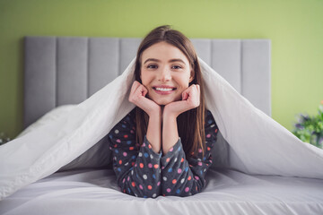Young brunette woman smiling while lying in bed under a duvet, dressed in polka dot pajamas,...