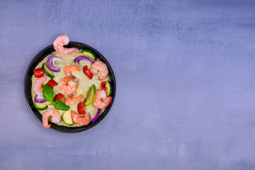 A plate of shrimp and vegetables in a black plate on a gray background. The creamy texture of the sauce is garnished with red onion rings, zucchini slices, red pepper pieces and fresh basil