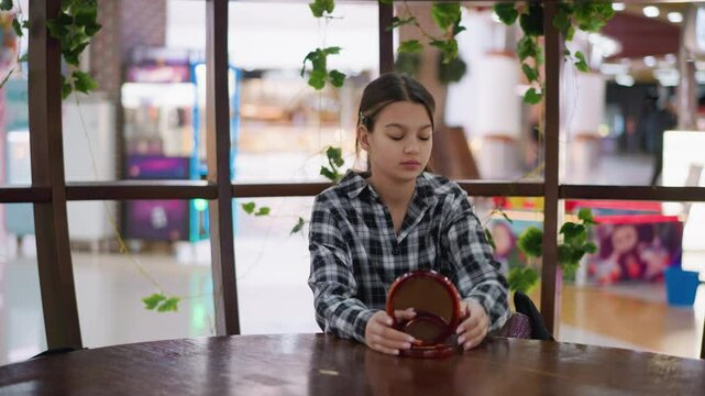 Teen girl in checkered shirt removes wooden mirror and two purses from her bag, placing the mirror upright and opening a purple purse while sitting in a cozy indoor space with plants and warm lights