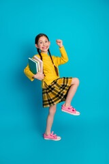 Smiling schoolgirl in yellow plaid skirt holding book, posing energetically with a colorful backpack