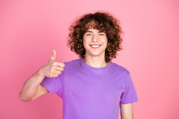 Smiling young man in a purple t-shirt showing a thumbs-up gesture against a pink background,...