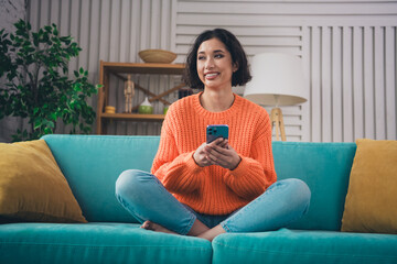 Young woman in a cozy orange sweater smiling while holding a phone, sitting cross-legged on a teal...