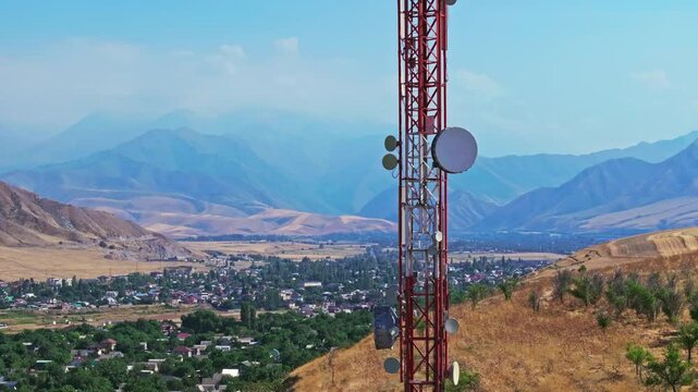 Drone footage showing a mountain village near with a telecom tower on a dry hill. The drone flying slowly upwards in front of the tower, creating a parallax effect with distant mountains in the