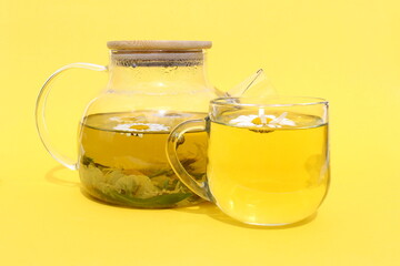 On a yellow background there is a glass teapot with a cup of chamomile infusion.