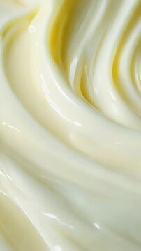 Creamy white icing being slowly swirled and textured in macro view. The soft, glossy frosting creates gentle flowing patterns as it's shaped, with subtle yellow tones appearing throughout the motion.