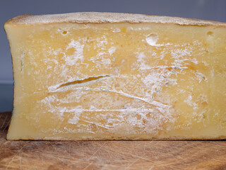 a large piece of mature cheese with a hard, natural rind. The cheese has a yellowish color, irregular holes and traces of mold on the surface. It lies on a wooden board
