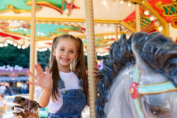 Happy emotional child riding on a merry go round at amusement park. Little girl playing on carousel, summer fun, happy childhood, vacation and entertainment concept