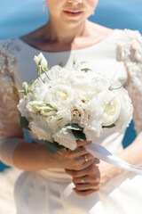 bride is holding a bouquet of white flowers. The bouquet is large and has a lot of flowers in it. The woman is wearing a white dress and a gold ring on her finger. Scene is elegant and romantic.