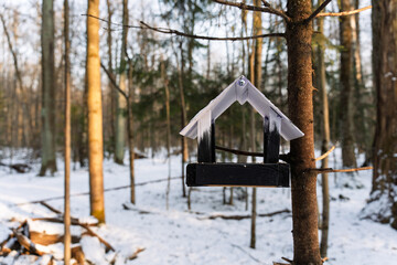 A handmade wooden bird feeder painted in black and white hangs on a tree in a snowy winter forest. The sun casts warm light through the bare trees, creating a peaceful and atmospheric scene.