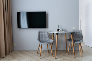A modern minimalist interior with a small round table and two gray chairs against a light gray wall. A flat-screen TV is mounted on the wall, and a decorative reed diffuser is placed on the table.