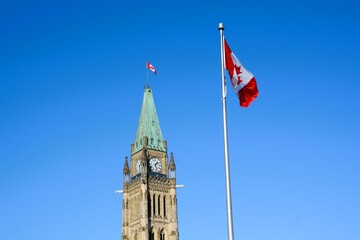 The Canadian flag flies against a clear blue sky with the top of the majestic Peace Tower of the Canadian Parliament Buildings in the background in Ottawa, Ontario, Canada.