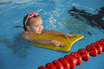 Little girl with goggles and kickboard swimming in pool indoors