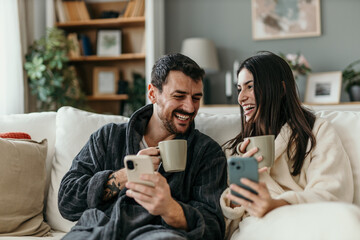 Couple wearing bathrobes laughing and drinking coffee while using smartphones on sofa