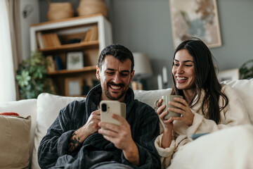 Couple Wearing Bathrobes Relaxing on Sofa Using Smartphone and Drinking Tea